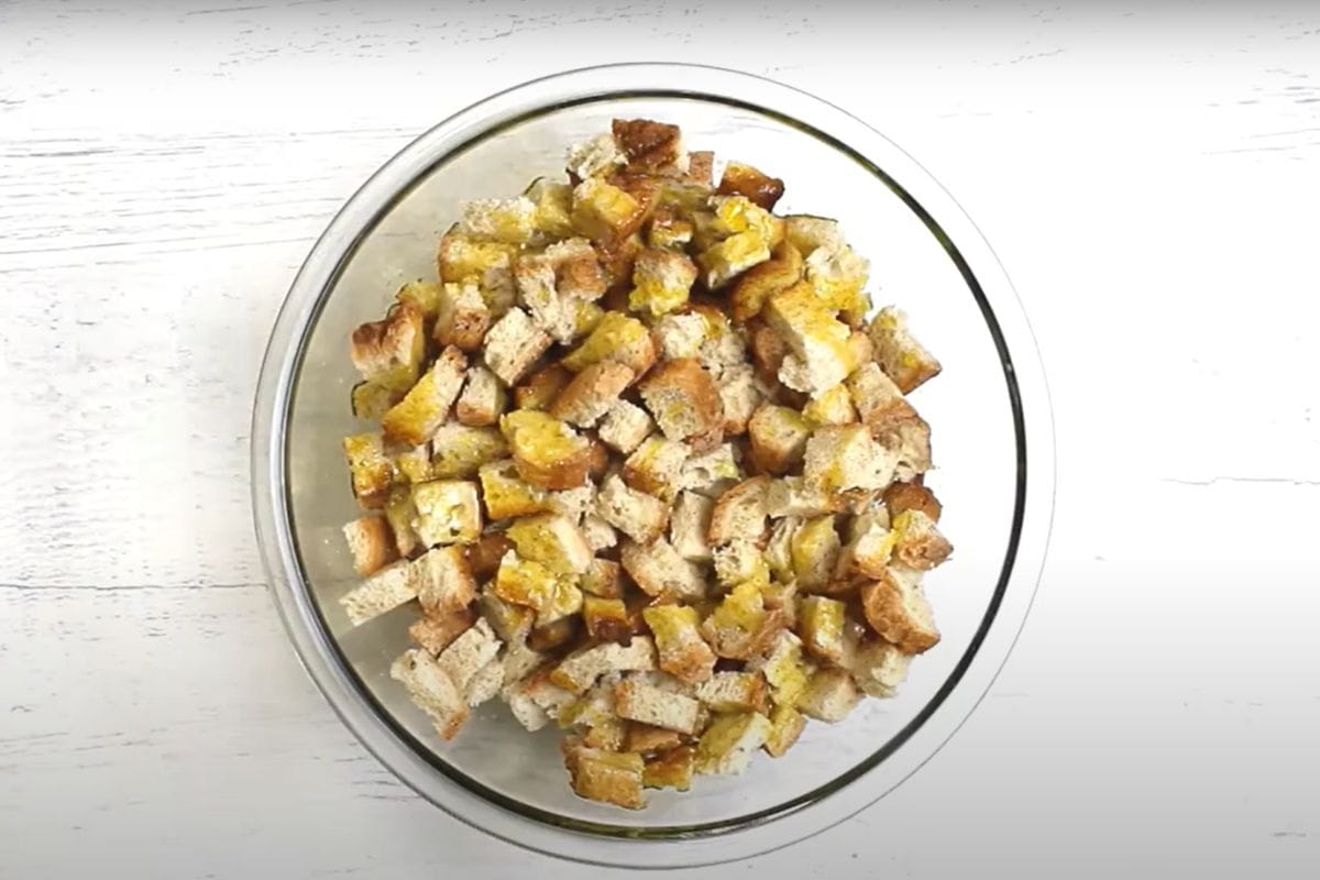 Gluten-free bread cubes covered in olive oil to make croutons in a glass mixing bowl.