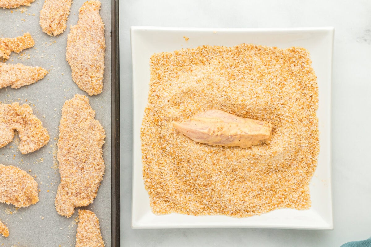 tossing a chicken tender in panko breadcrumbs on a white plate.