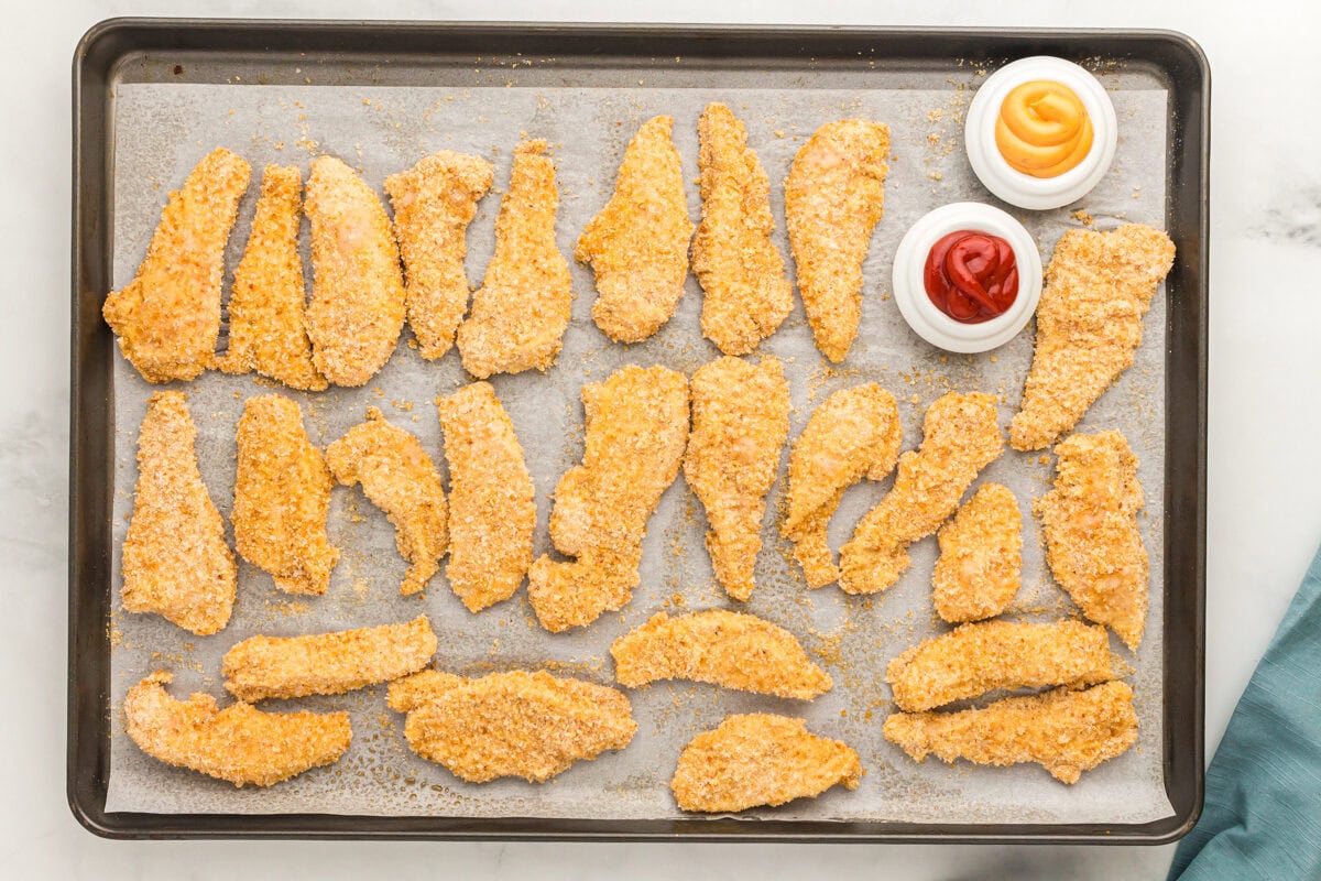 baked gluten-free chicken tenders on a baking sheet with ketchup and mustard.