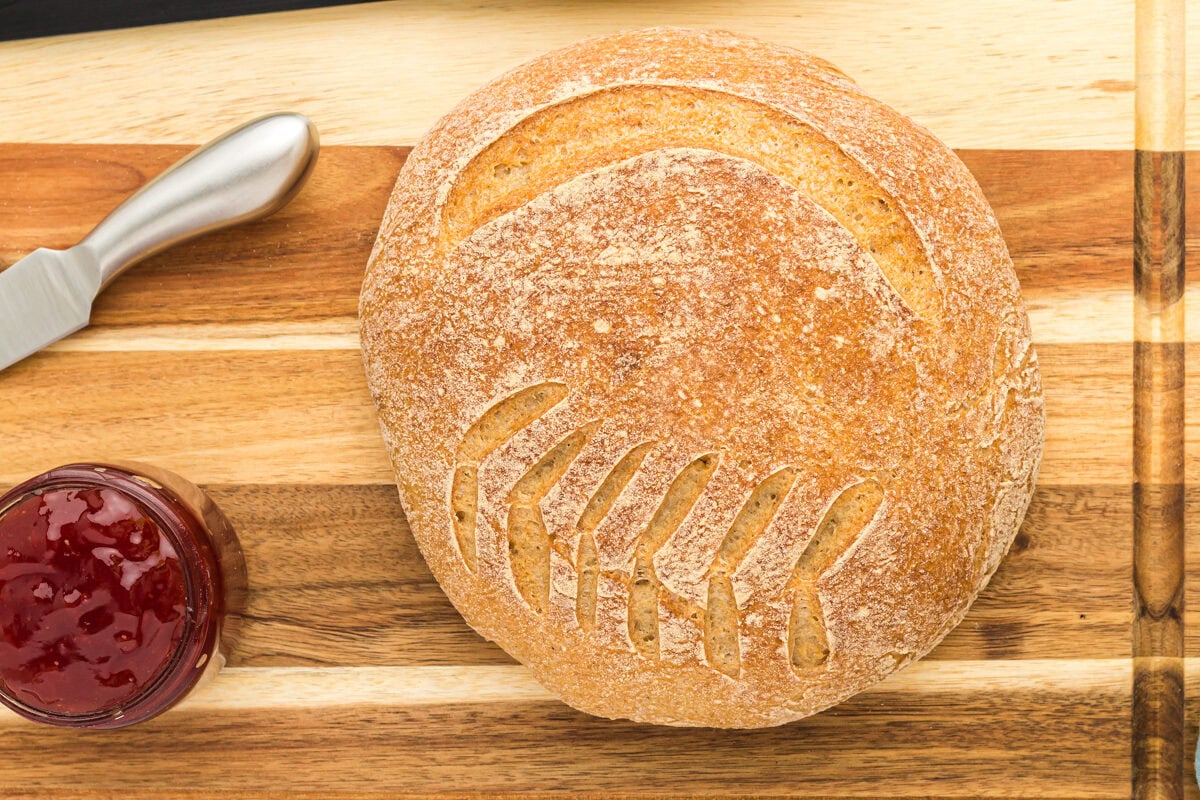 close up of a boule of gluten free sourdough bread on a wooden cutting board with a knife and jam.