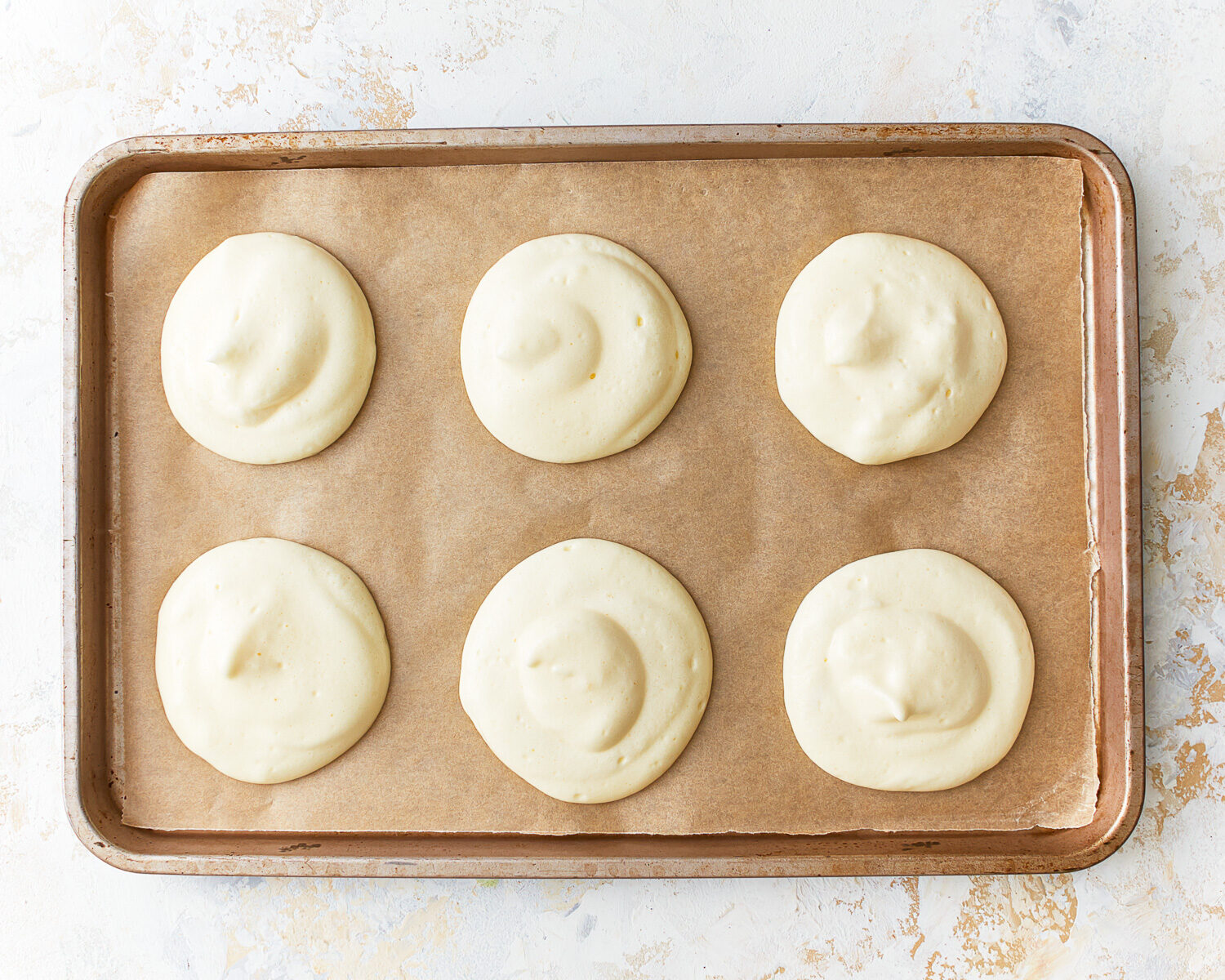 Six rounds of cloud bread batter on a parchment lined baking sheet.