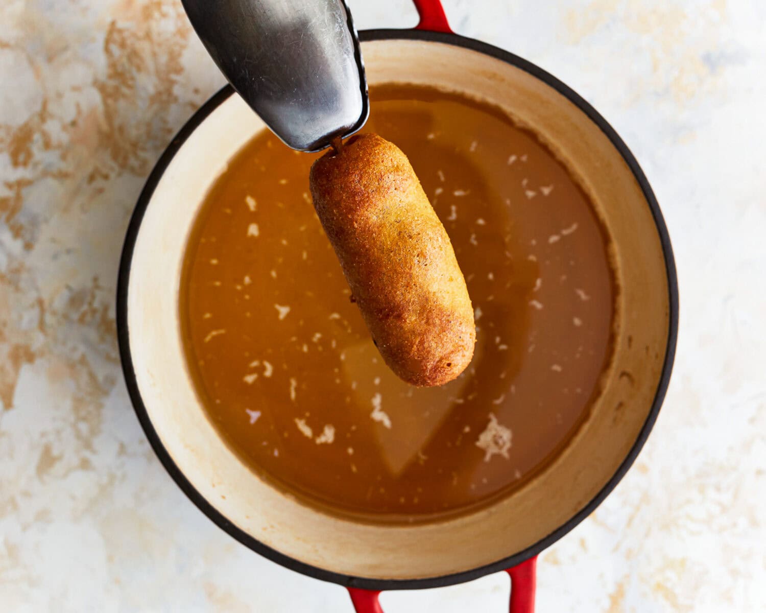 holding a cooked corn dog over a pot of oil with tongs.