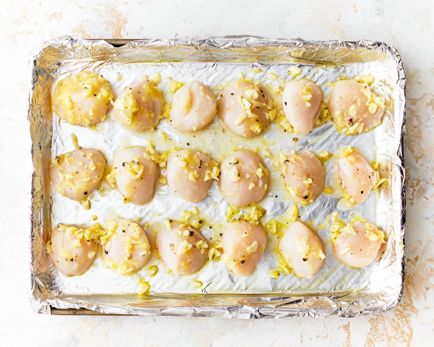 Overhead view of raw scallops on a foil lined baking sheet covered in garlic butter.