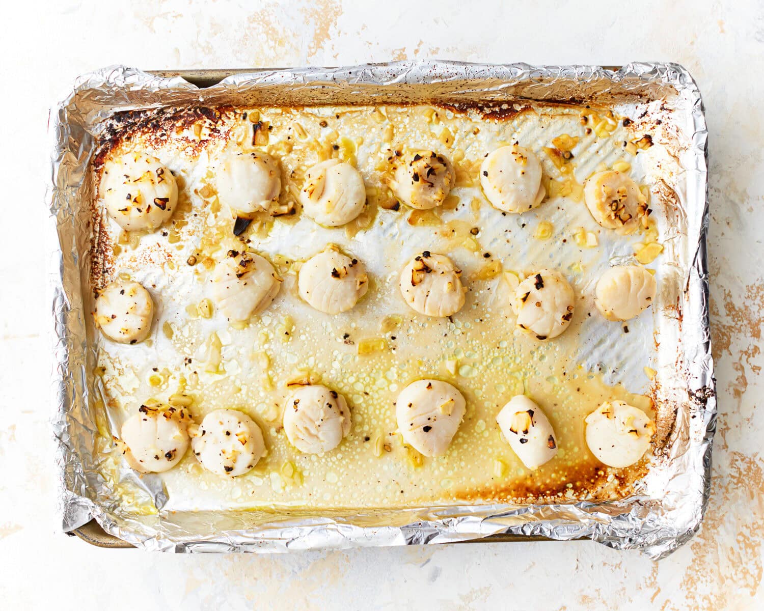 Overhead view of broiled scallops on a foil lined baking sheet covered in garlic butter.