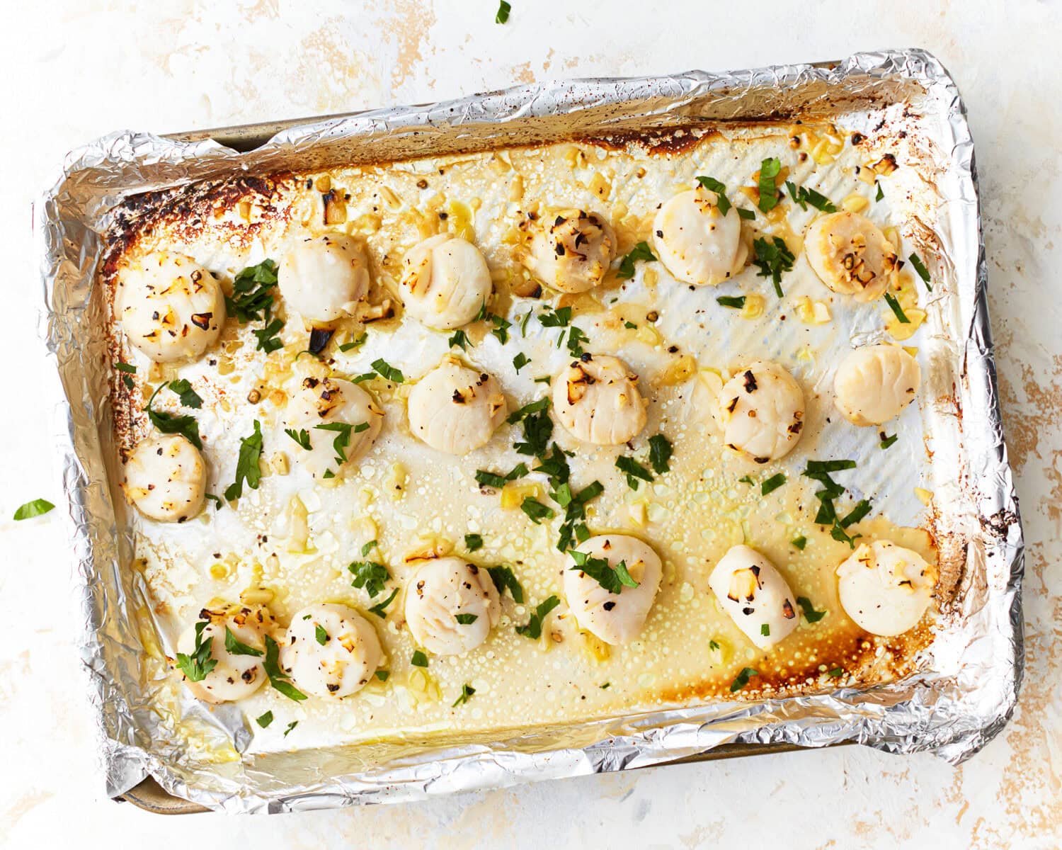 Overhead view of broiled scallops on a foil lined baking sheet covered in garlic butter and parsley.