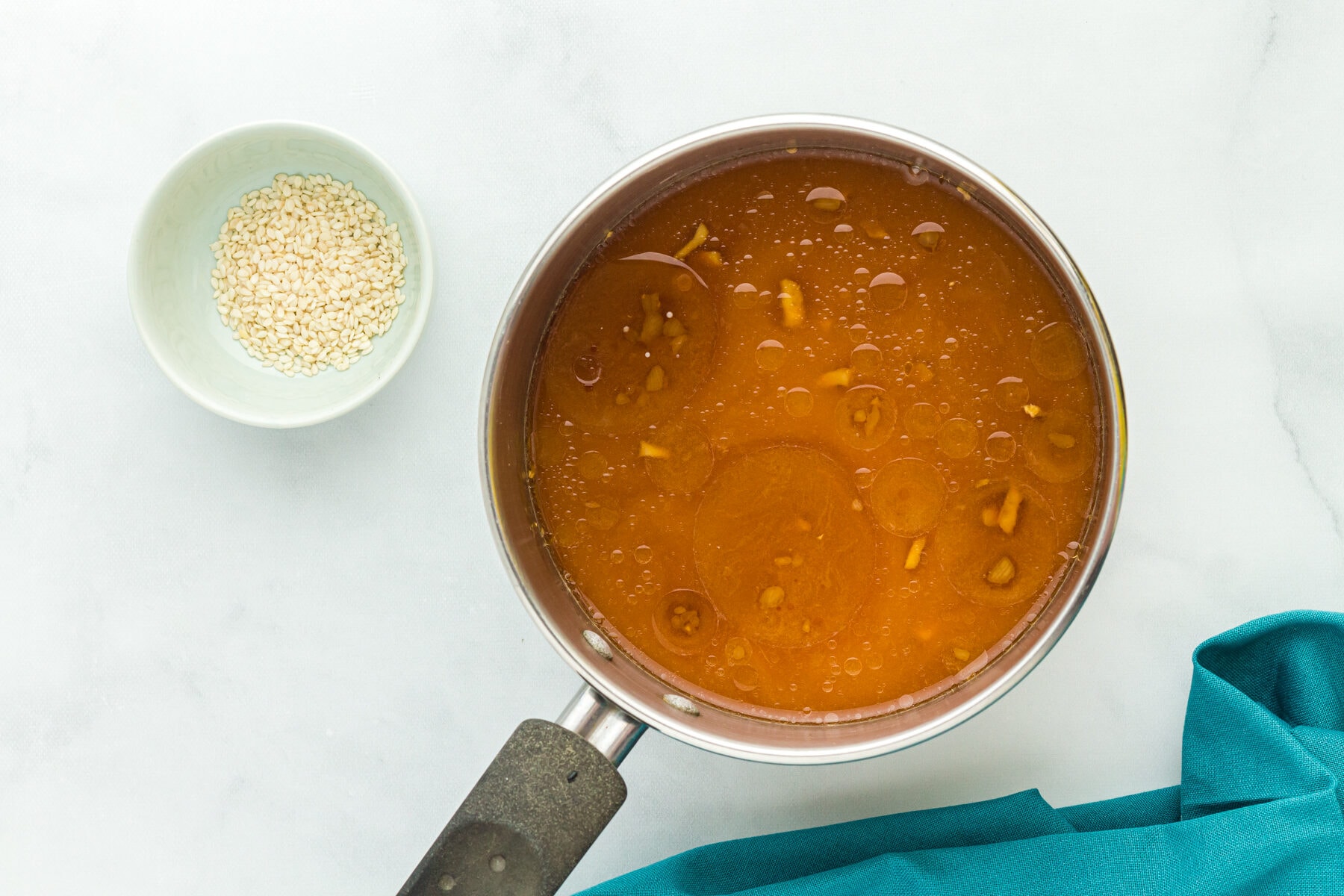 Overhead view of cornstarch slurry mixed into gluten free teriyaki sauce in a saucepan next to a bowl of sesame seeds.