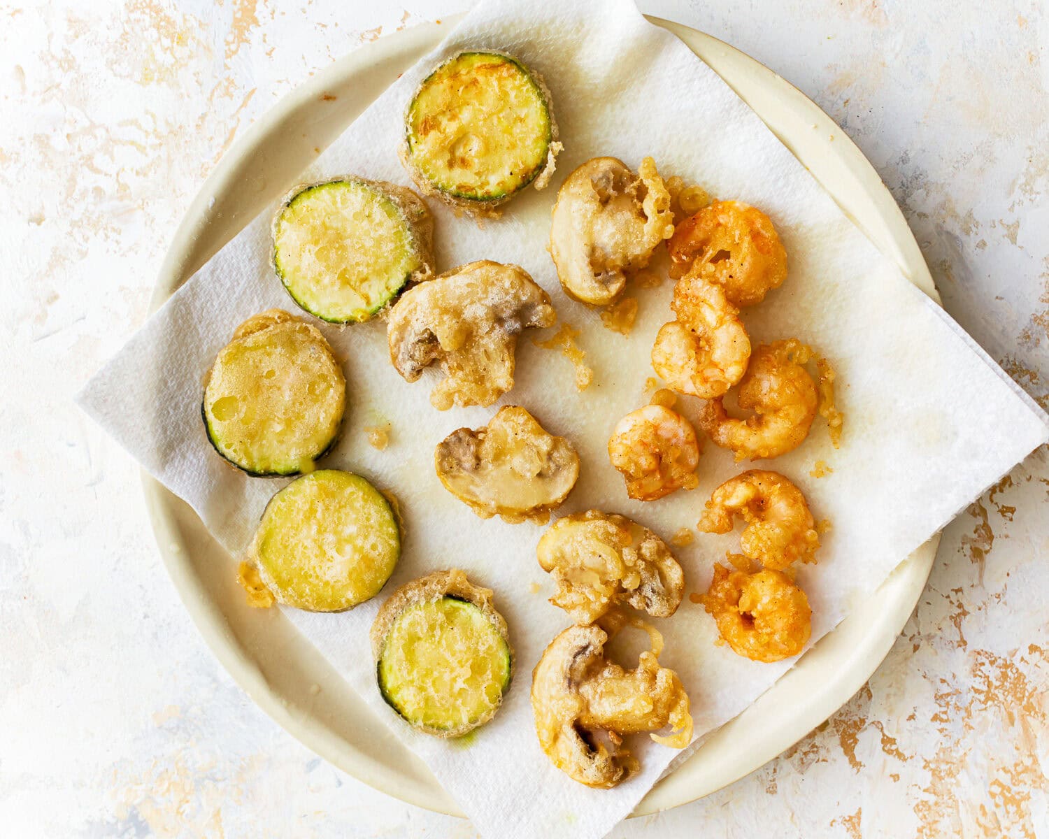 Vegetables and shrimp cooked in a gluten free tempura batter on a paper towel lined plate.