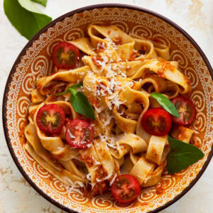 A close up of a bowl of almond flour pasta with fresh vegetables, shredded cheese, and herbs