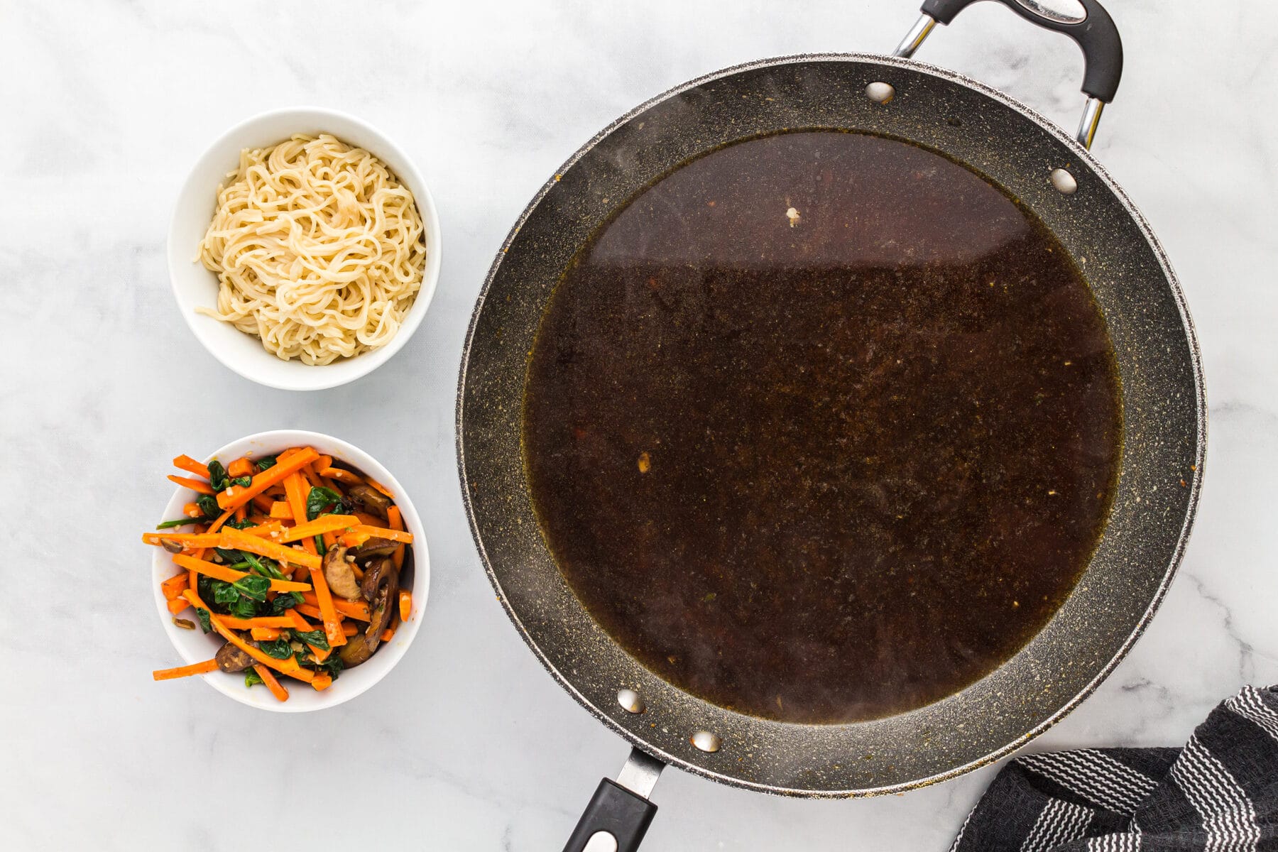 Chicken broth in a skillet next to a bowl of vegetables and gluten-free ramen noodles