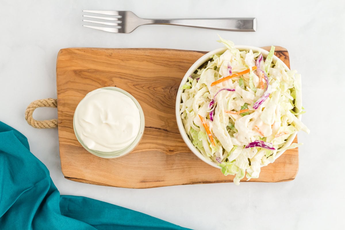 A small pot of vegan mayonnaise next to a bowl of coleslaw on a wooden chopping board