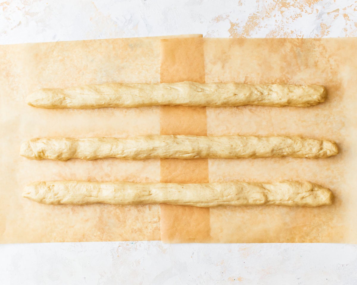 Gluten-free challah bread dough divided into three and rolled into a long rope shape on parchment paper.